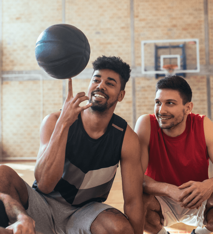 Young basketball player spinning a ball on his finger – he and his friend are smiling as they sit on the bench courtside