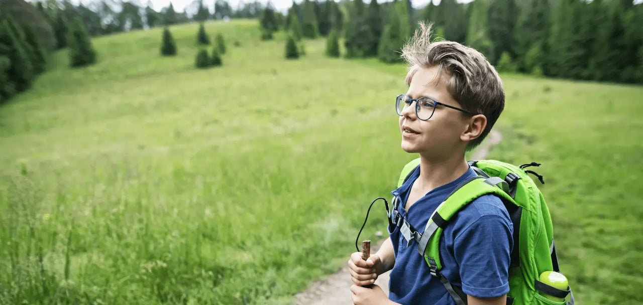 Young boy on a hike across a meadow with pine trees in the distance. It’s an adventure and
he’s carrying a small backpack