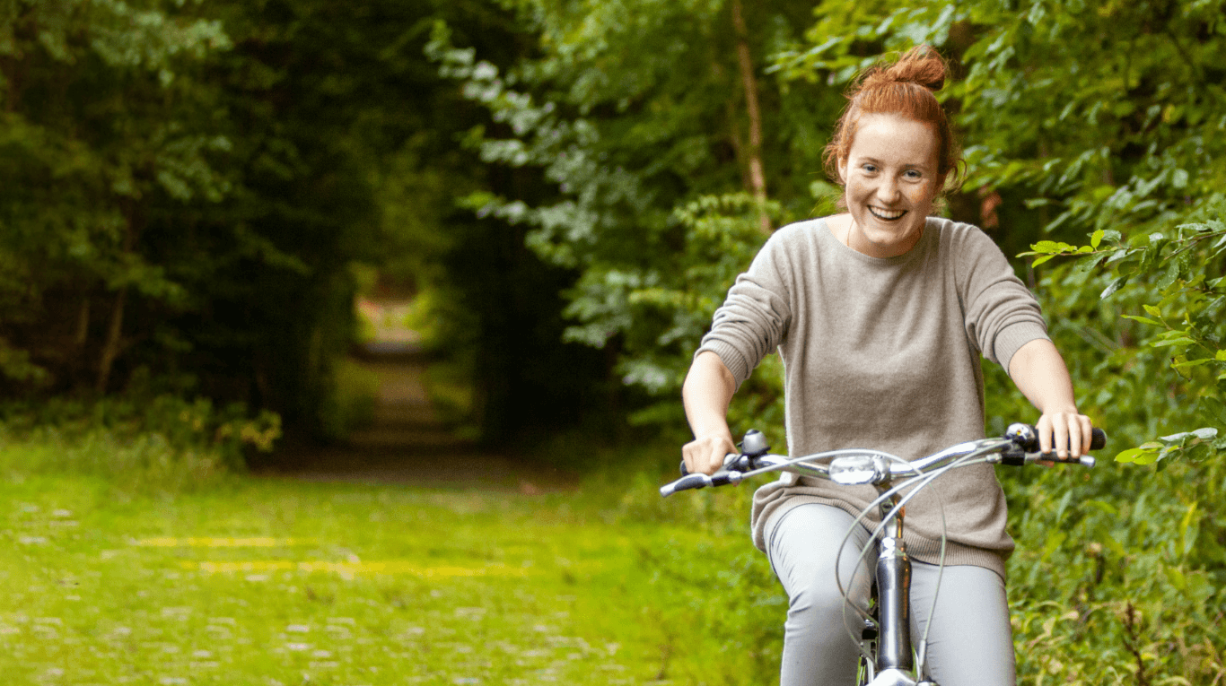 Happy young woman riding her bicycle towards the camera – it’s a bright spring day and everything is looking very green